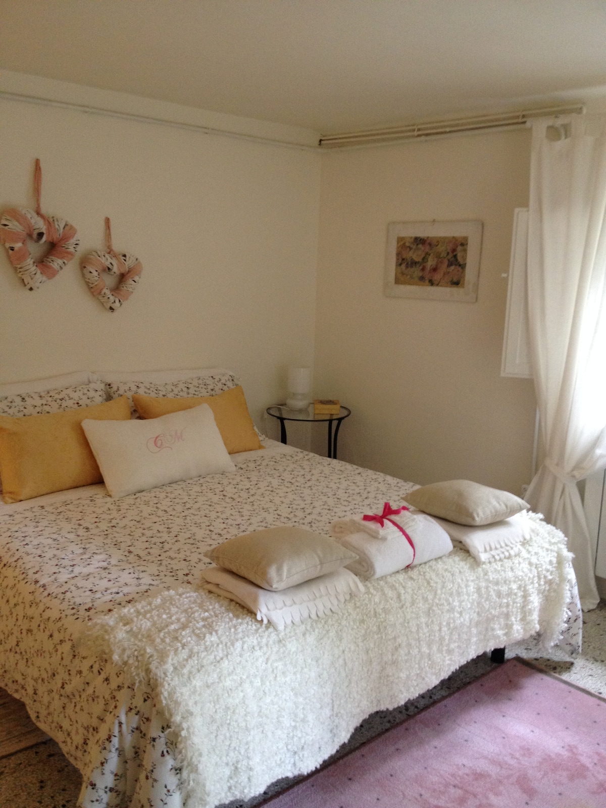 A welcoming sleeping area features a spacious bed adorned with light-colored linens and decorative pillows. Soft natural light enters through a window dressed with sheer curtains. A small side table holds a lamp and other items, adding to the room's cozy ambiance.