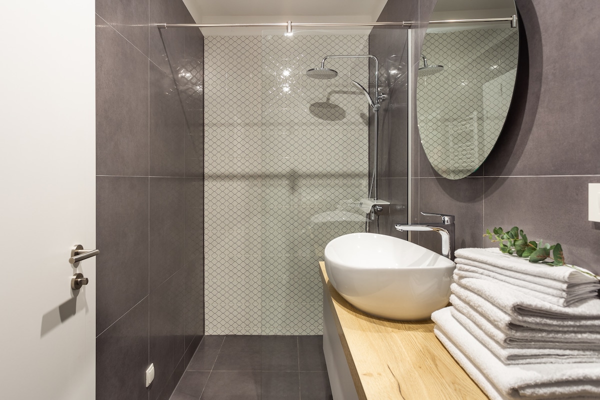 A modern bathroom is featured, highlighted by a walk-in shower with a glass partition and stylish tile design. A circular mirror above a wooden countertop complements a white basin. Neatly arranged towels are stacked on the countertop, enhancing the clean aesthetic.