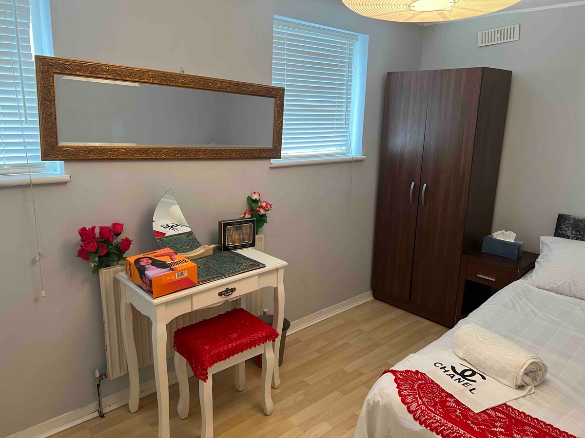 A well-lit bedroom features a spacious bed with white linens and a decorative red throw. A wooden wardrobe stands against the wall, complemented by a vanity table topped with a mirror and a few decorative items. Fresh flowers are placed nearby for a touch of elegance.