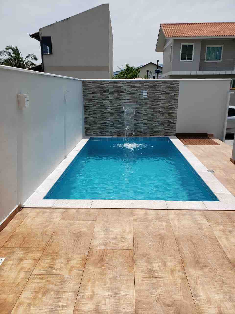 A swimming pool is depicted, featuring clear blue water and a relaxing waterfall feature. The surrounding area includes a tiled deck, and the walls are adorned with a textured stone design. Sunlight illuminates the space, creating a welcoming outdoor atmosphere.