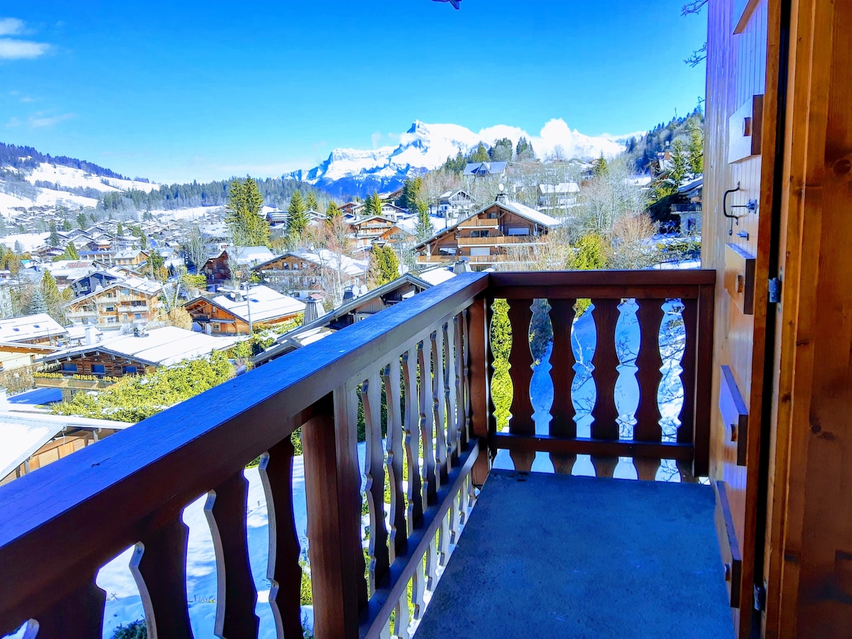 A balcony is visible, framed by wooden railings, overlooking a scenic view of snow-covered mountains and village rooftops. Clear blue skies enhance the serene atmosphere, while details of nature and architecture can be appreciated in the distance.