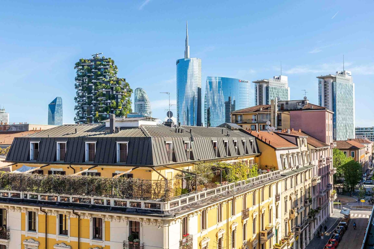 A panoramic view from the apartment reveals the modern skyline of Milan, showcasing the towering buildings of the financial district. The foreground features a charming residential building with a traditional facade and lush greenery along the terrace.
