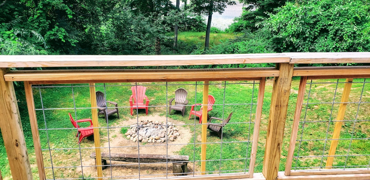 A fire pit area is encircled by colorful Adirondack chairs, creating a welcoming space for gatherings. The pit is surrounded by a stone ring, with a log placed for additional seating. Lush greenery and trees provide a natural backdrop, reflecting the tranquil surroundings.