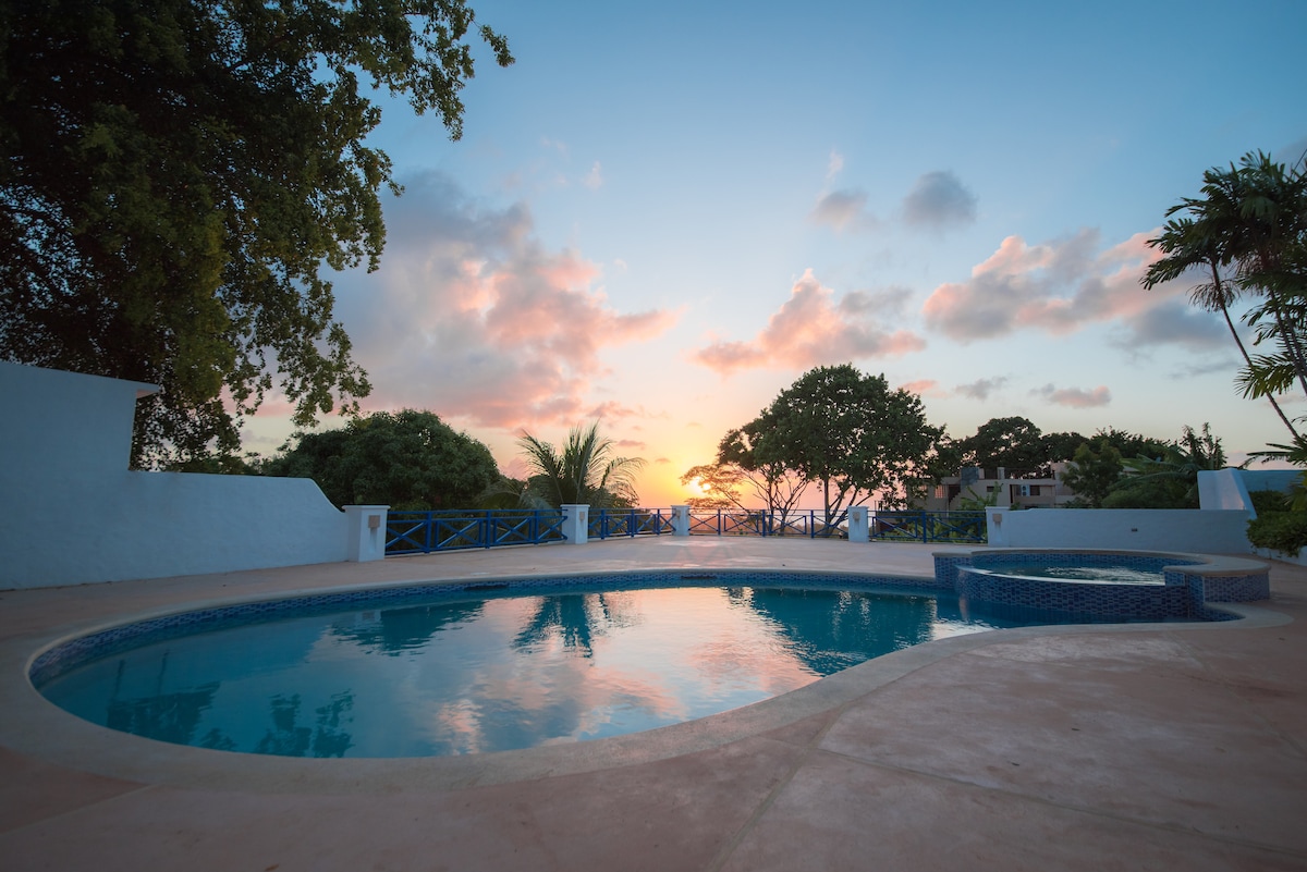 A tranquil pool area is set against a colorful sunset sky, highlighting the serene water surface. Lush greenery and scenic trees frame the space, while blue railings and a small spa are visible nearby, creating a peaceful outdoor setting.