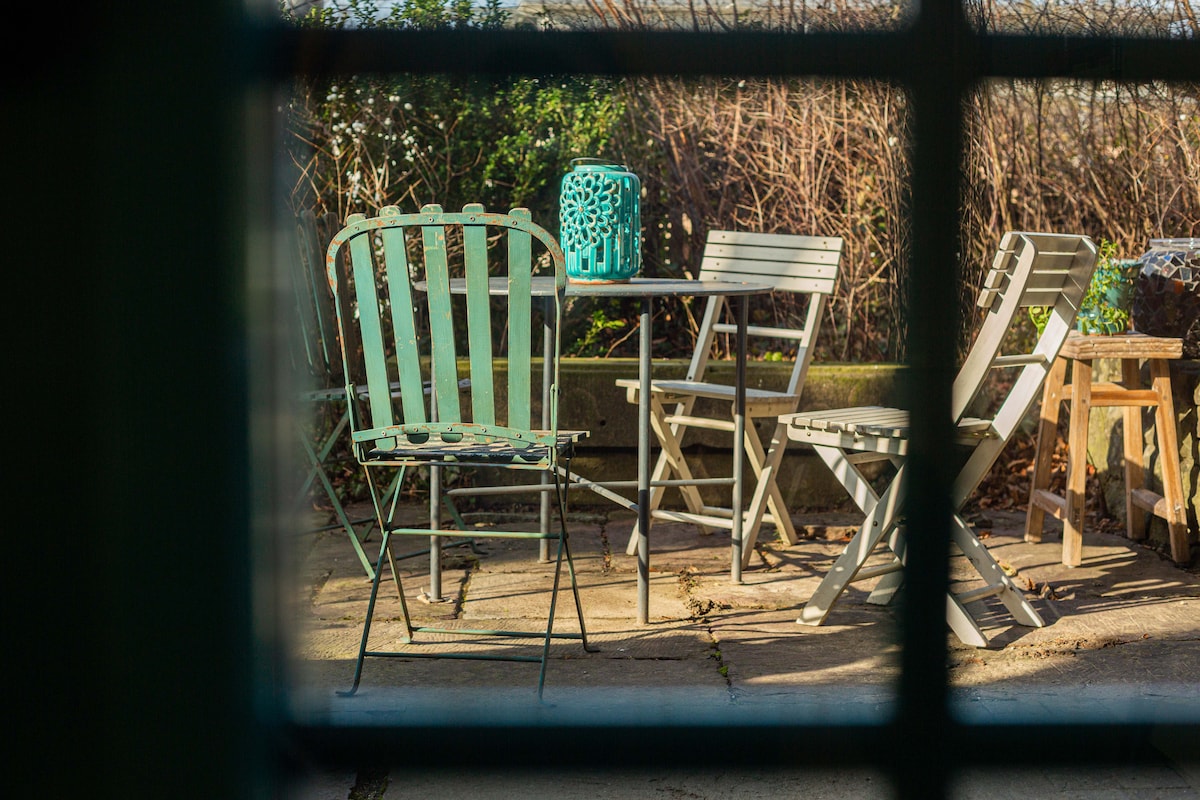 A private patio area is visible through a window frame, featuring a mix of outdoor furniture including a green metal chair, a wooden table, and light-colored folding chairs. The surrounding greenery provides a natural backdrop, creating a serene outdoor space.