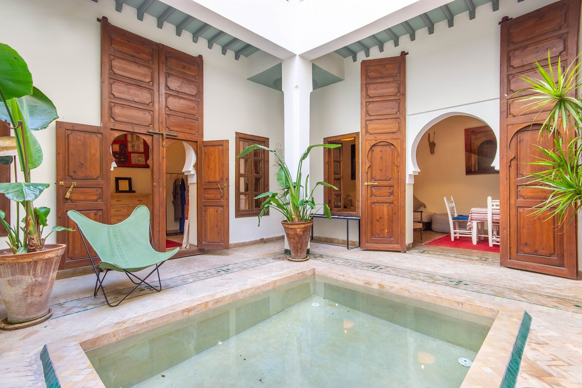 A traditional courtyard is surrounded by wooden doors and large potted plants, creating a serene atmosphere. A small, shallow pool is centered in the space, reflecting the bright light from above. Comfortable seating is positioned nearby, inviting relaxation.