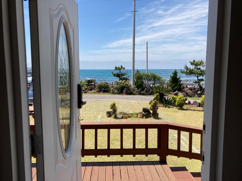An open front door reveals a scenic view of the ocean, with lush greenery and a well-manicured lawn in the foreground. Delicate shrubs are positioned along the pathway, while a clear blue sky enhances the tranquil atmosphere.