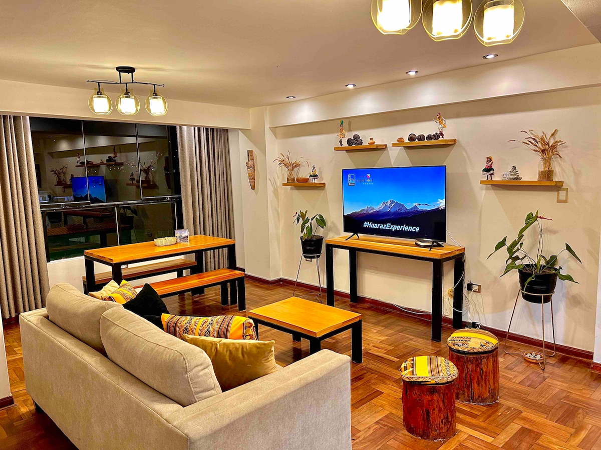 A modern living area features a comfortable sofa with decorative cushions, complemented by wooden tables and natural accent pieces. A television is mounted on the wall, while large windows allow natural light to fill the space. Indoor plants add a refreshing touch.