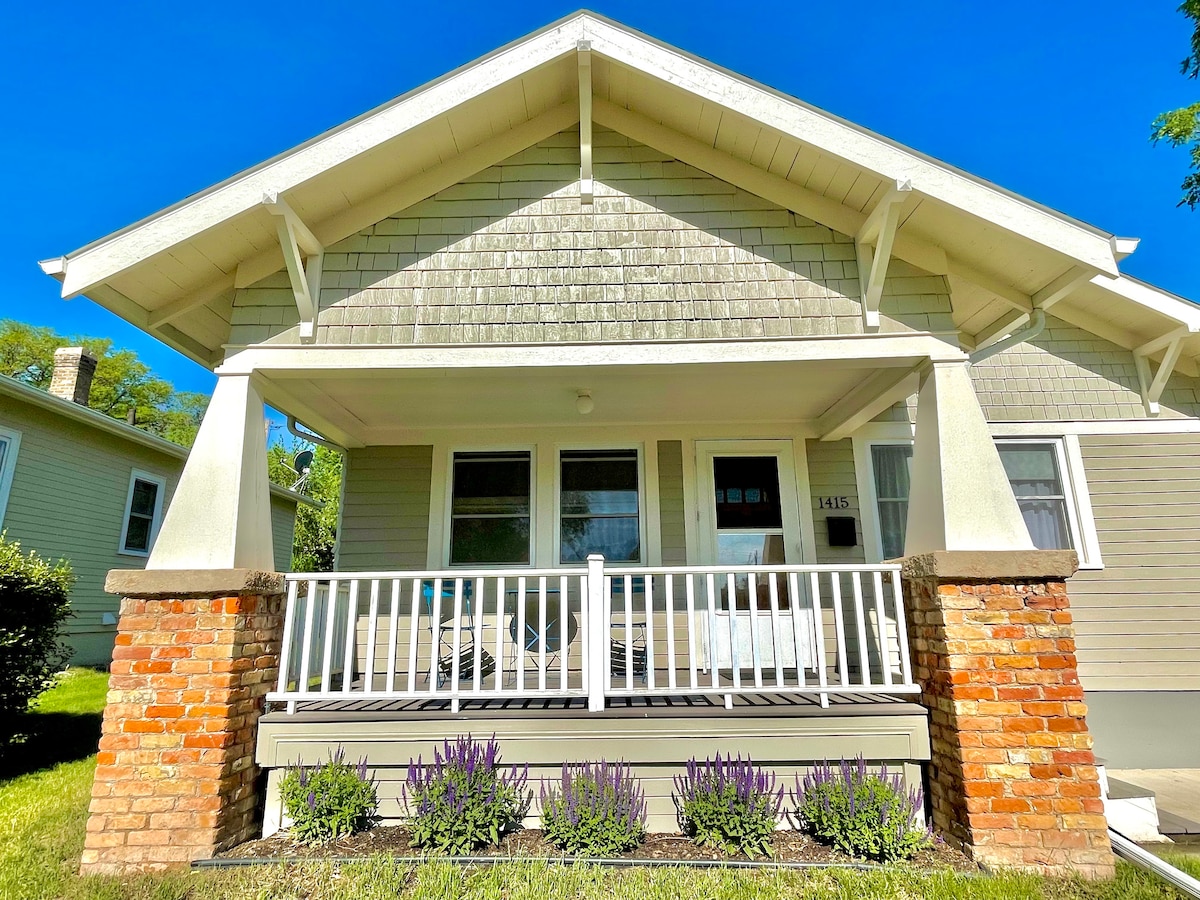 A charming front porch is showcased with a wooden railing and comfortable seating. The porch is flanked by brick pillars, and well-maintained lavender plants are visible along the front. The home features a light-colored exterior and a sloping roof under a clear blue sky.