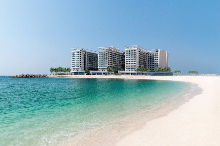 A modern beachside building is set against a clear blue sky, featuring multiple stories and balconies. The structure is surrounded by soft white sand and the calm turquoise waters of the Arabian Gulf, creating a serene coastal environment.