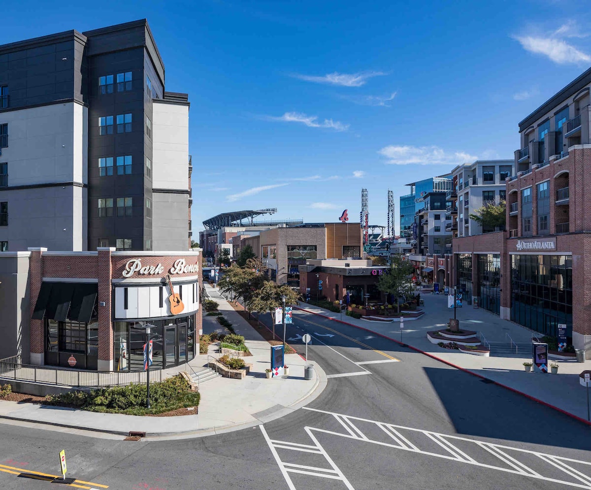 The image captures a vibrant urban scene featuring modern buildings, including a restaurant with a prominent sign reading 'Park Bench'. The photo showcases wide sidewalks, street lamps, and clear blue skies, emphasizing the accessibility of nearby Truist Park and local attractions.