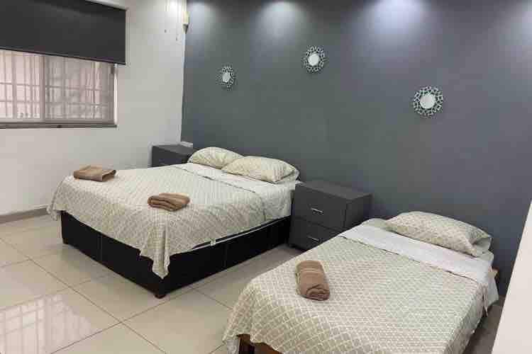 A well-lit sleeping area is presented with two beds covered by light-patterned linens. Each bed is accompanied by neatly arranged towels. A minimalist grey wall is enhanced by circular wall mirrors, and a window offers natural light, partially covered by a roll-up shade.