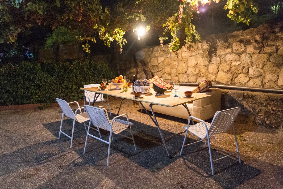 An outdoor dining area features a long table set with plates and glasses under softly glowing lights. Surrounding the table are several lightweight chairs. A stone wall and lush greenery provide a natural backdrop, enhancing the space’s inviting ambiance.