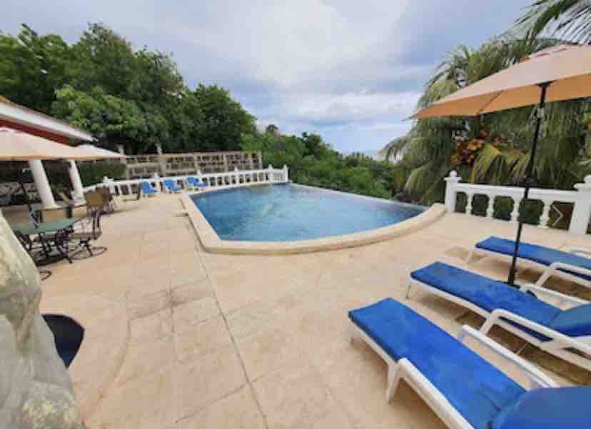The outdoor area features a spacious pool surrounded by a sun deck with lounge chairs, offering vibrant blue cushions. Parasols provide shade, and a dining table sits nearby under a covered patio. Lush greenery is visible around the pool area, enhancing the serene environment.