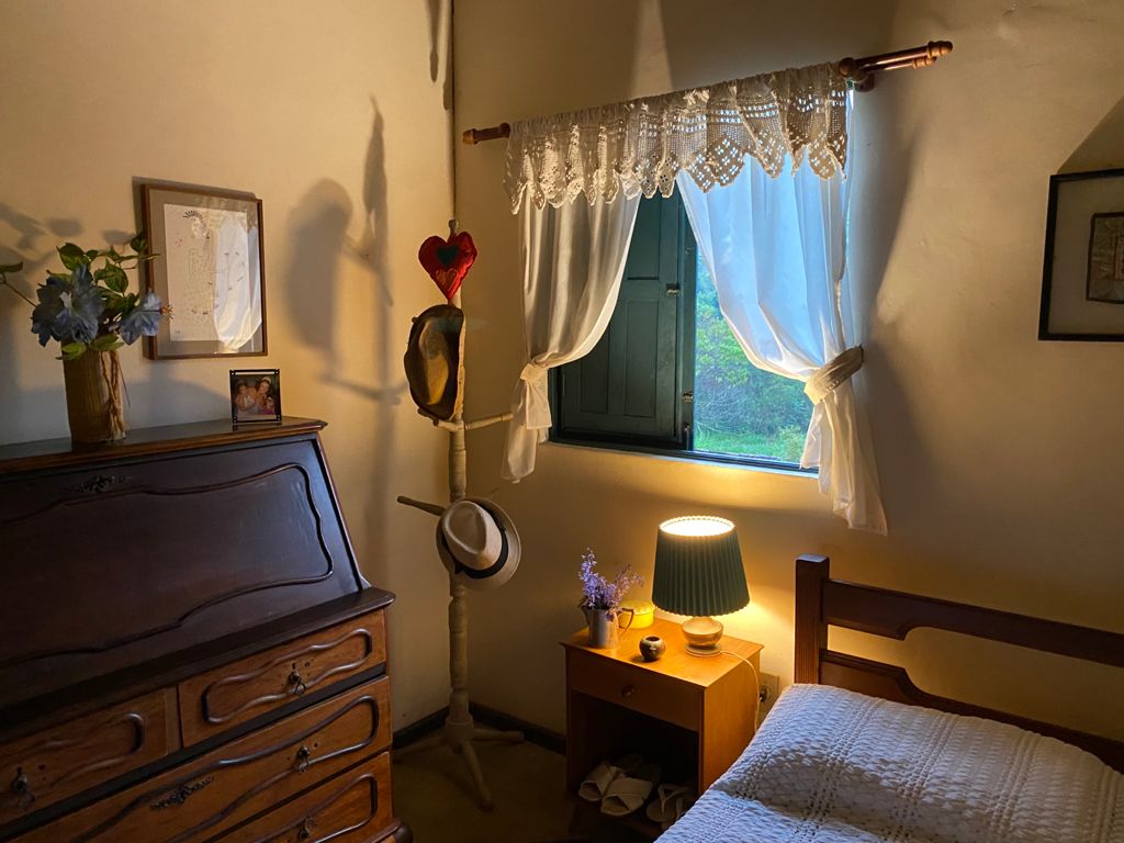 A cozy bedroom features a spacious bed beside a wooden nightstand with a lamp. A dresser with intricate details is placed against the wall. Natural light enters through a curtained window, highlighting the room’s warm tones. A decorative hat stand is positioned nearby.