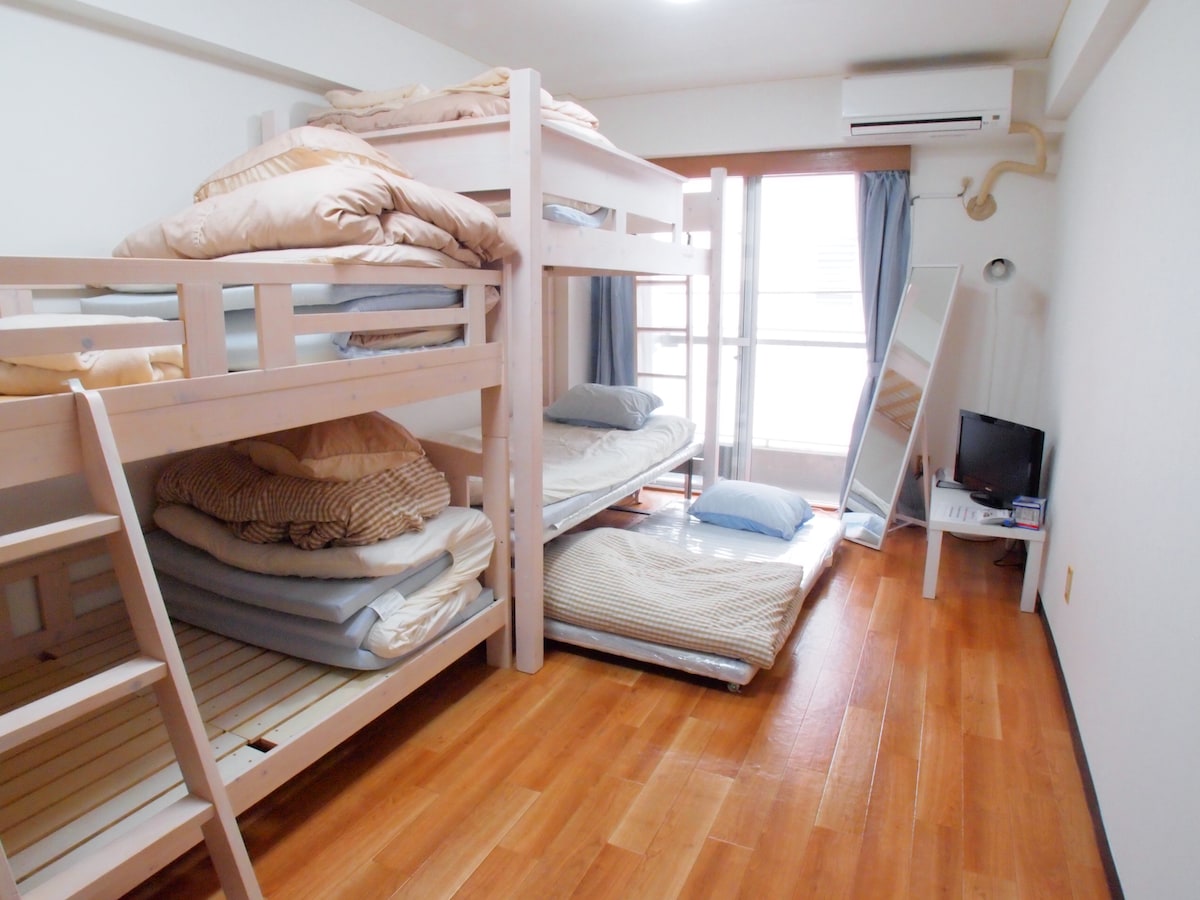The room features two sets of bunk beds with neatly arranged bedding. A large window allows natural light to fill the space, which is complemented by hardwood flooring. A TV and a mirror are positioned against the wall, enhancing the room's functionality.