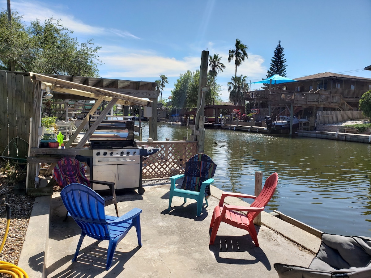 An outdoor patio area is visible by the canal, featuring a grill and several colorful chairs arranged around a concrete surface. Lush greenery and nearby palm trees enhance the scenic view of the water, with boats docked along the shoreline in the background.