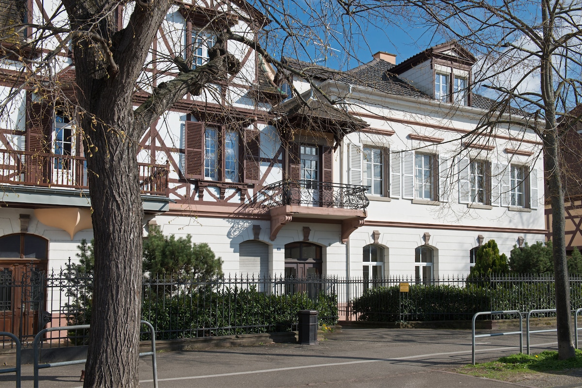 The historic building showcases a classic architectural style, featuring a mix of white and wooden facades. Large windows are framed by decorative accents, and a balcony extends from the upper level. Lush greenery and a wrought-iron fence are present in the front yard.