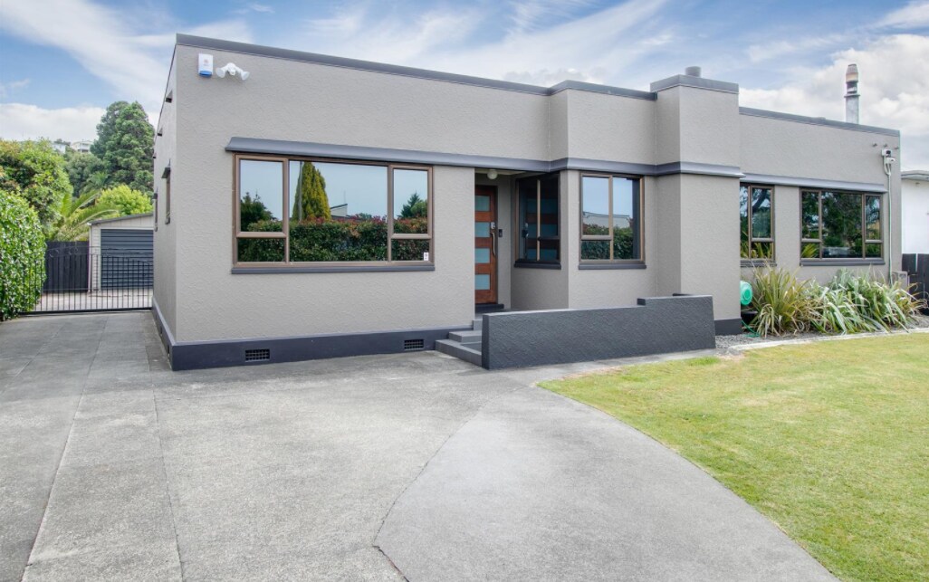 The exterior of the Art Deco house is shown, featuring a smooth grey facade with large windows. A curved driveway provides access to the entrance. Lush greenery surrounds the property, enhancing the peaceful atmosphere of the private yard, which is perfect for relaxation.