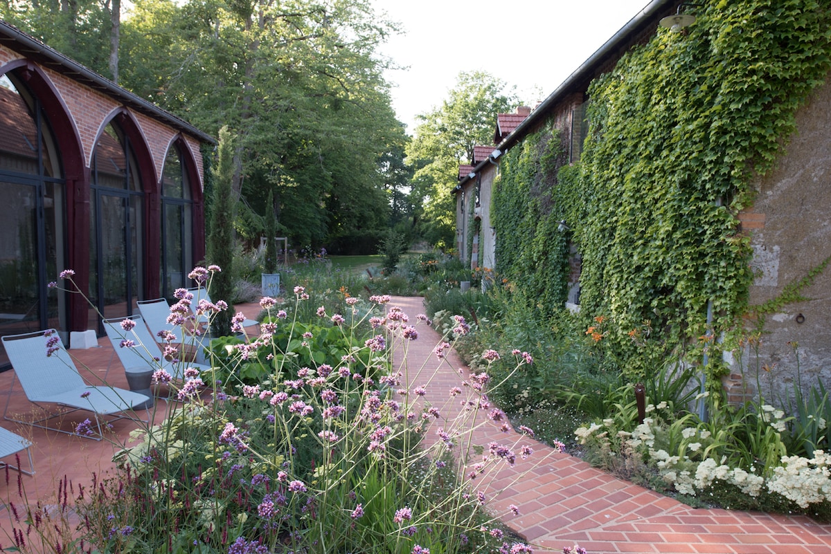 A winding brick path is surrounded by lush gardens, showcasing a variety of flowering plants. The path leads to a building with a stone facade, partially covered in greenery. Relaxing lounge chairs are positioned nearby, providing a serene space to enjoy the natural scenery.