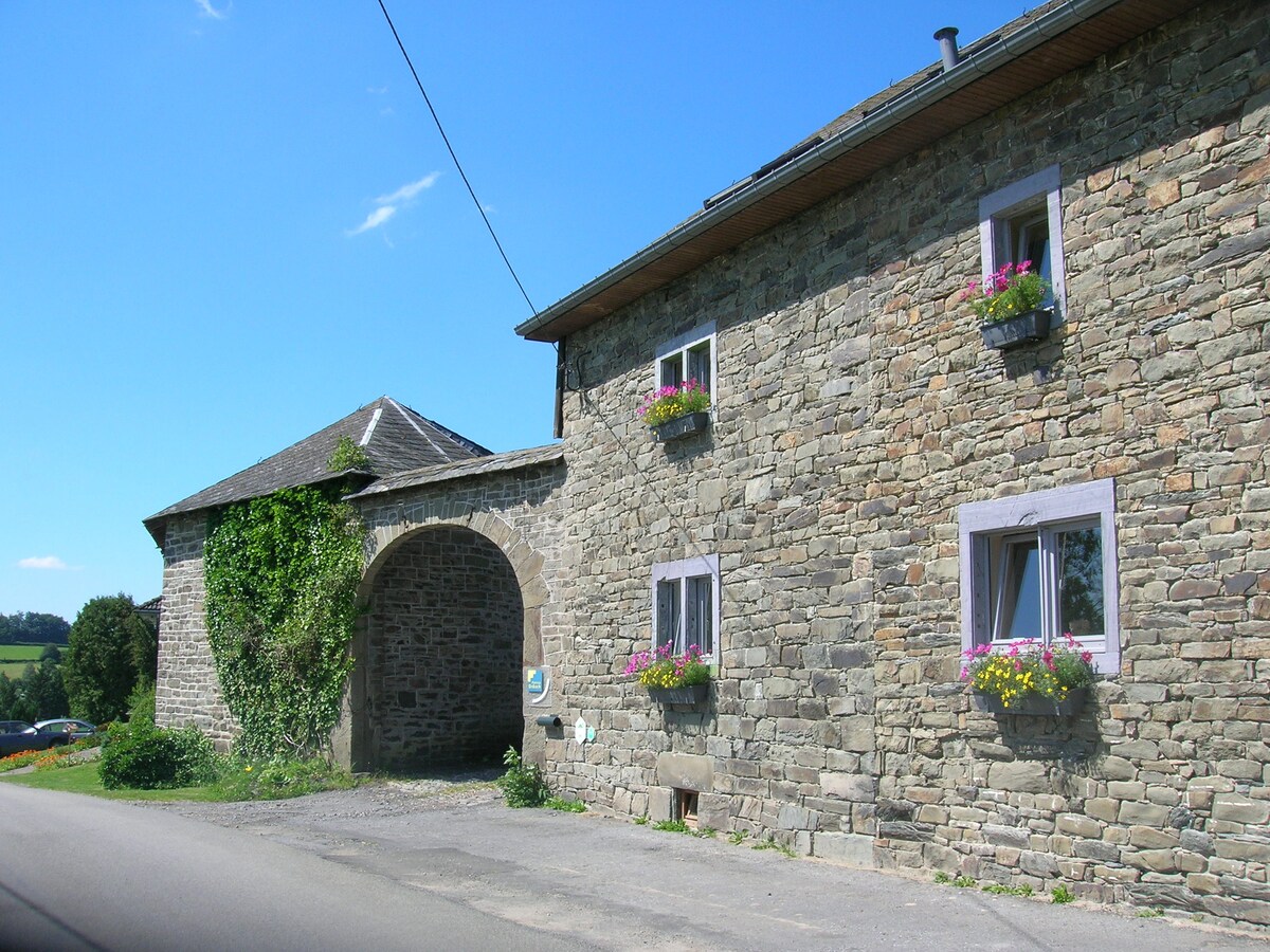 An old stone farmhouse features a distinctive archway along with several windows adorned with colorful flower boxes. The building is surrounded by greenery, and the clear blue sky adds to the serene rural setting.