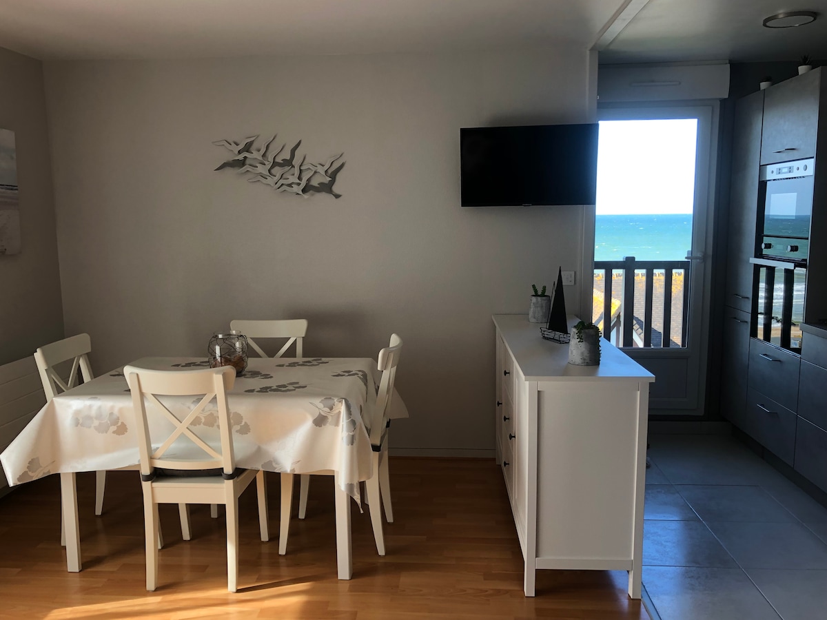 A dining area is visible, featuring a light wood table surrounded by white chairs. A television is mounted on the wall, with a sideboard offering storage. Large windows bring in natural light and provide views of the sea.