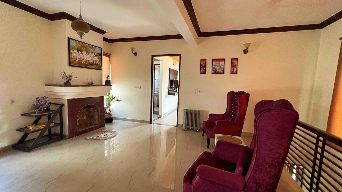 A comfortable living area features two plush armchairs in deep purple, positioned near a cozy fireplace. Decorative wall art and small plants add a touch of charm, while sunlight filters through the windows, illuminating the tiled floor and enhancing the welcoming environment.