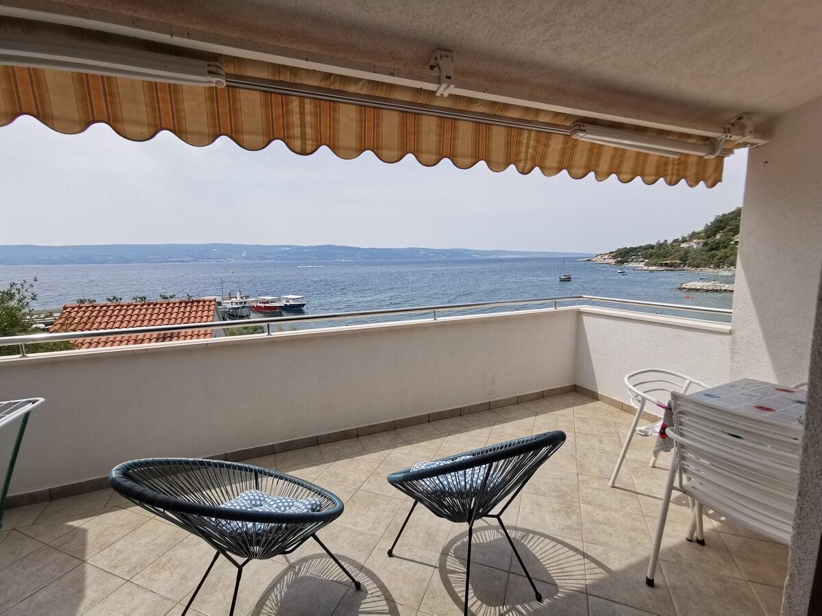 A spacious terrace offers stunning views of the sea, featuring two wicker chairs and a table, with a retractable awning providing shade. The tiled floor complements the serene coastal scenery, where the horizon meets gentle waves.