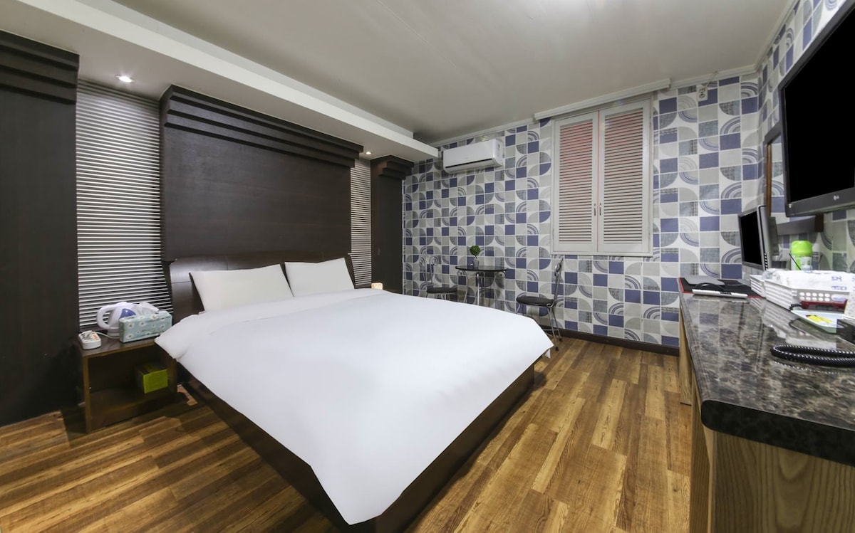 A spacious sleeping area features a large bed with crisp linens and simple decor. The walls present a geometric pattern, and natural light streams through a large window. A desk is positioned nearby, providing a functional workspace, while minimalist furniture adds to the room's efficiency.