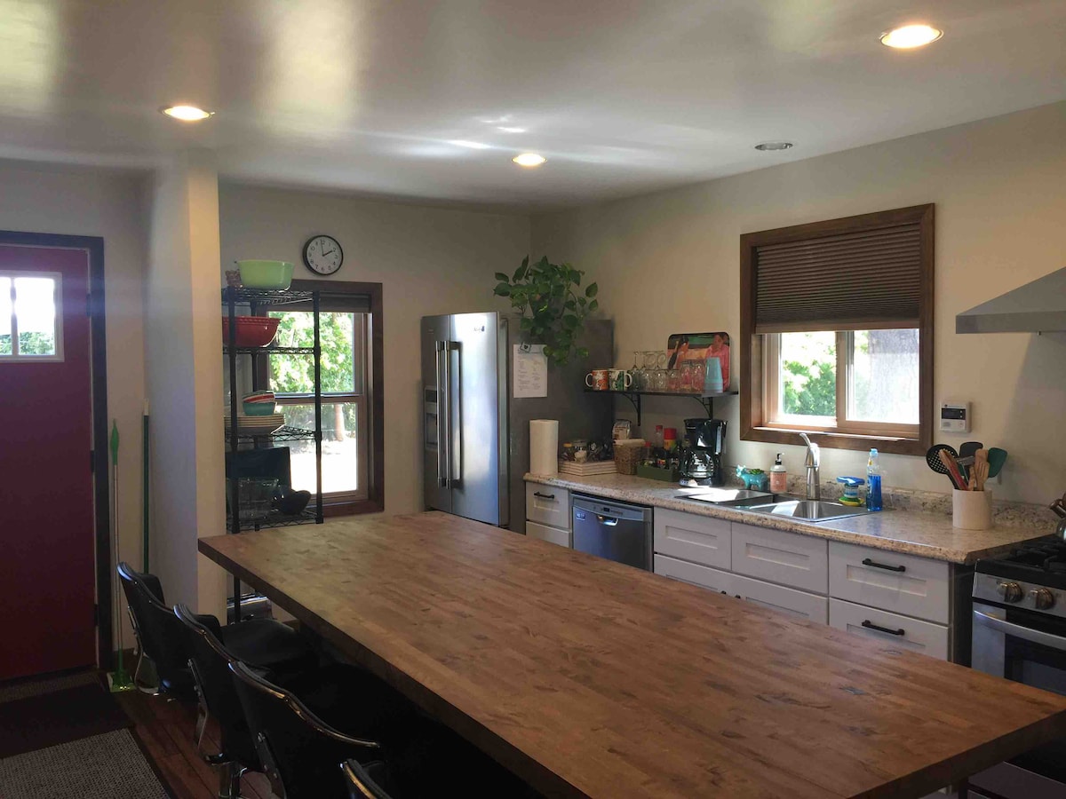 A modern kitchen features a large wooden island surrounded by four black chairs. Appliances include a stainless steel refrigerator and stove. Natural light enters through a window and a red door can be seen in the background, with plants adding a touch of greenery.
