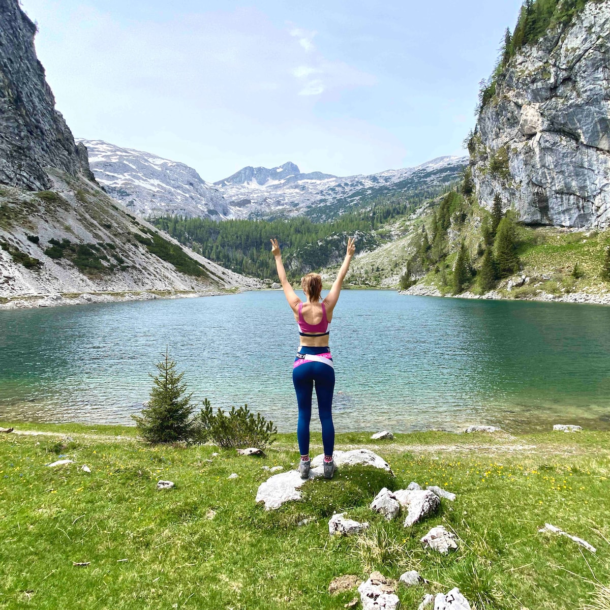 A person stands on a rock overlooking a serene lake surrounded by majestic mountains. The clear water reflects the rocky cliffs and lush greenery. The sky is partly cloudy, enhancing the natural beauty of the landscape.