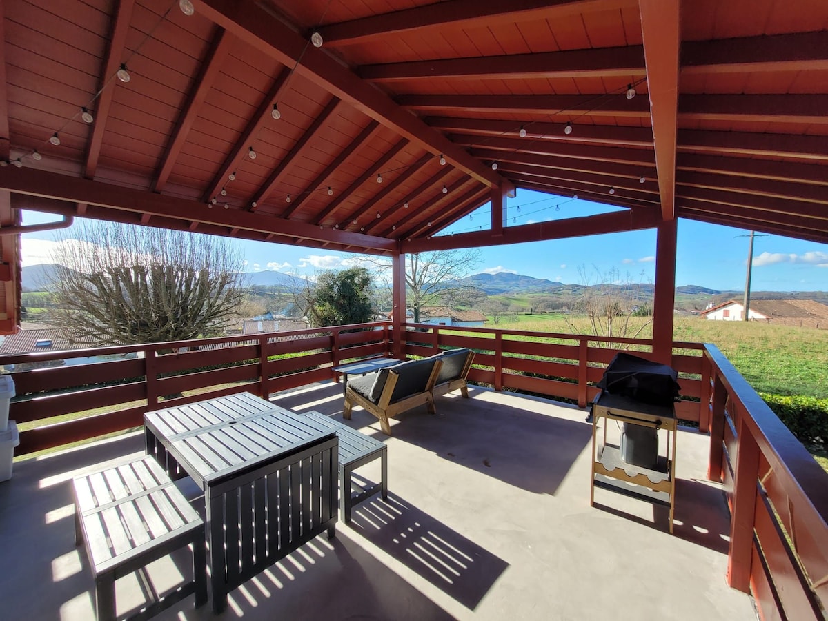 A spacious terrace is covered with a wooden roof, featuring a dining table with seating and two lounge chairs. Surrounding greenery and distant hills are visible, creating a serene outdoor space. String lights are hung along the beams, adding a cozy touch.