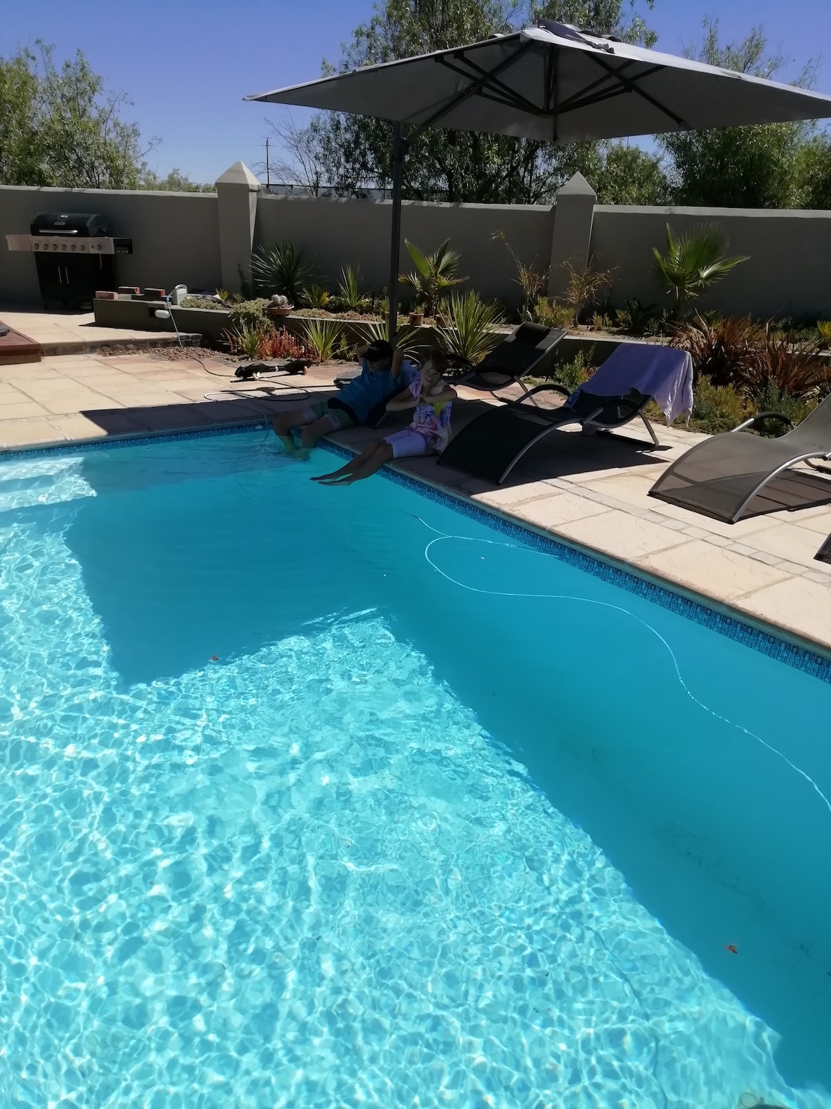 A refreshing pool is surrounded by lounge chairs and an umbrella, providing shade. A barbecue grill is positioned nearby, and landscaped greenery adds a natural touch. Two individuals can be seen enjoying the space, with the clear water sparkling in the sunlight.