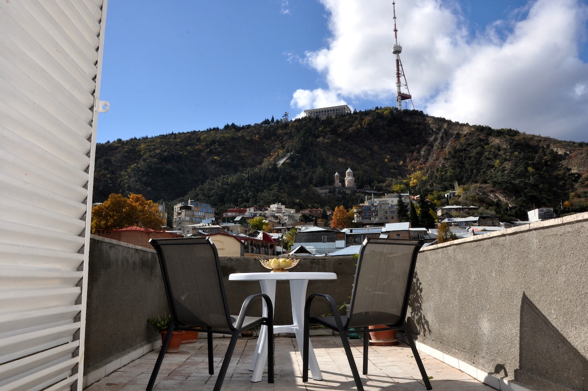 A private terrace is shown with two chairs and a small table, overlooking a hillside and the iconic television tower on Mtatsminda. The area is framed by gray walls, and the sky above features scattered clouds letting in light.