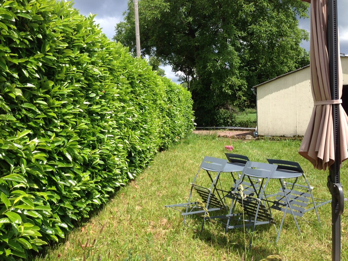 A peaceful garden space is visible, enclosed by tall greenery. A set of metal folding chairs is arranged on the grassy area, providing a spot for relaxation. The boundary features a structured hedge, contributing to a sense of privacy and tranquility.