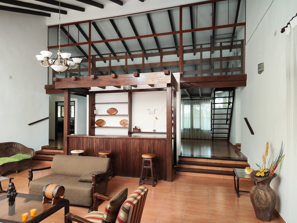 The spacious living area features high ceilings and a large wooden bar with decorative items on display. A cozy sofa set is arranged across from the bar, while a staircase leads to a lofted area above, enhancing the open feel of the space.