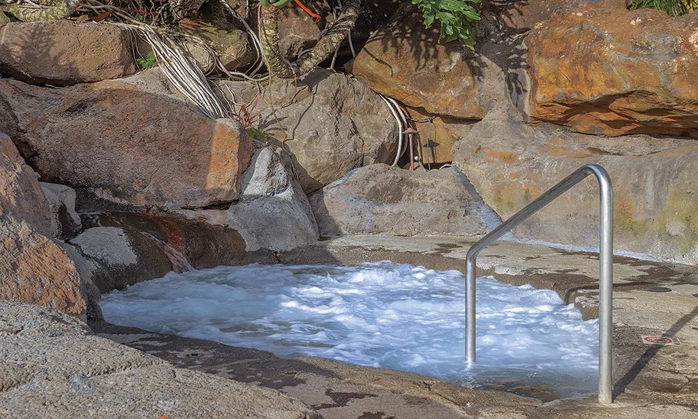 A secluded hot tub is surrounded by natural stone, with bubbling water creating a serene environment. The rock formations provide a rustic backdrop, while greenery partially frames the scene, contributing to the tranquil setting.