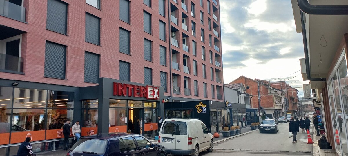 A modern building with a prominent red brick facade is visible, housing a store with large glass windows on the ground floor. The street is lined with various vehicles and pedestrians, surrounded by additional shops and buildings, illustrating an urban atmosphere.