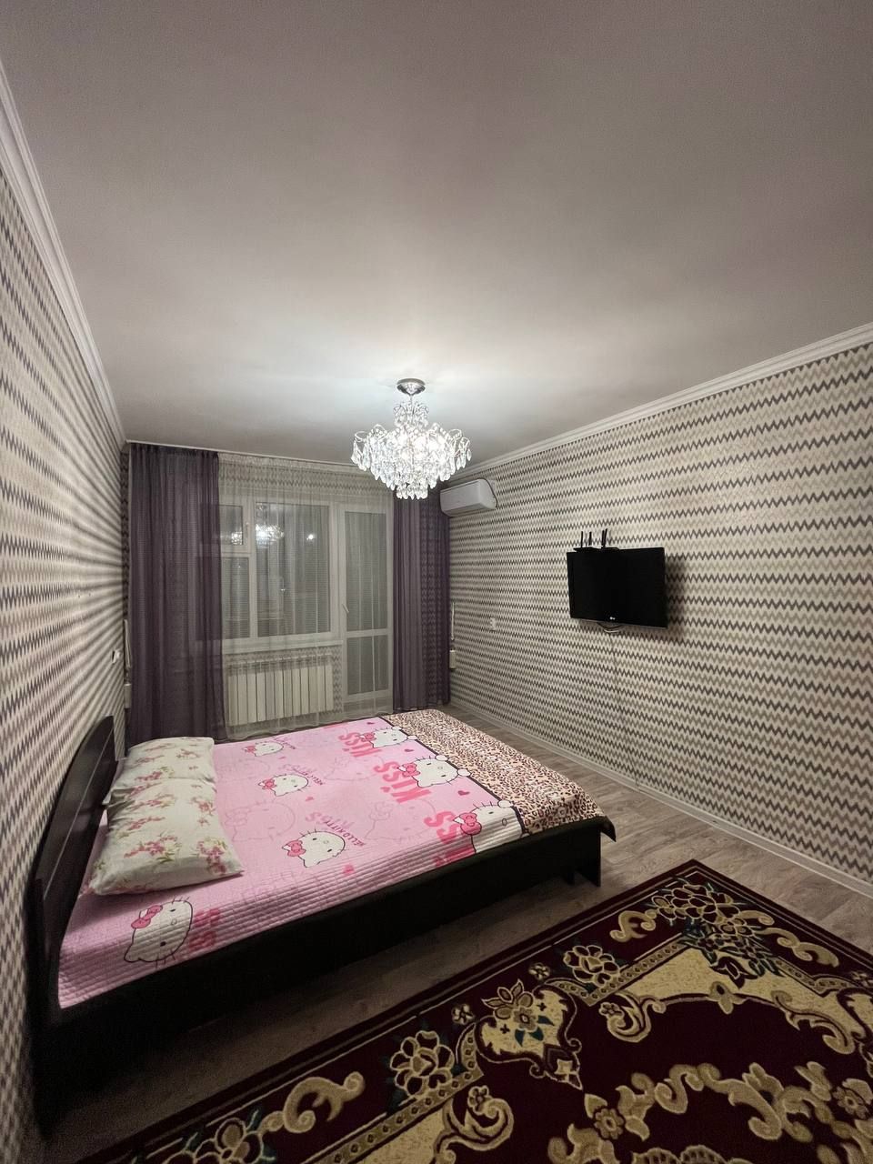 A cozy bedroom is equipped with a bed featuring a patterned pink blanket. Light streams through a window dressed with sheer curtains. A decorative chandelier hangs from the ceiling, while a television is mounted on the wall opposite the bed. A patterned area rug covers the floor.