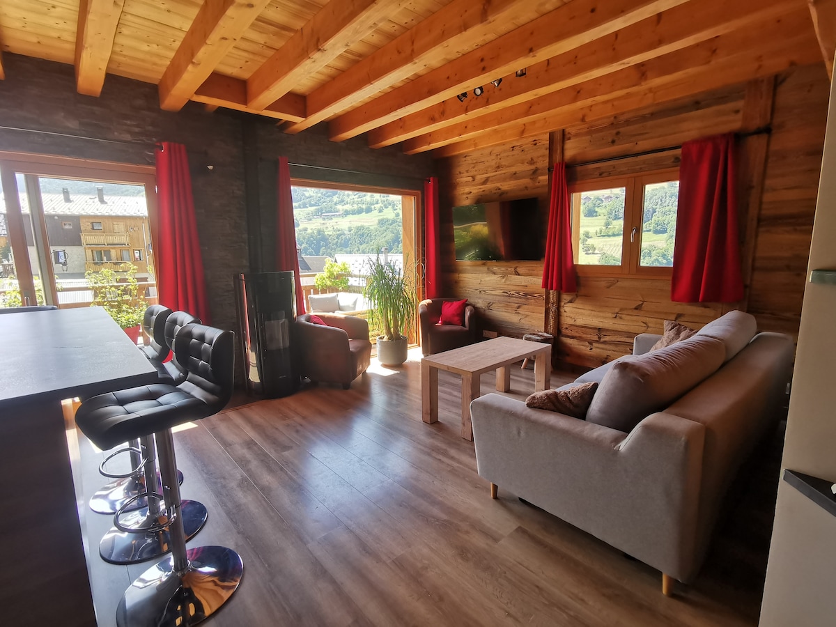 A spacious living area features large windows that invite natural light and provide views of the surrounding valley. Comfortable seating is arranged around a central coffee table. Red curtains frame the windows, and modern bar stools are positioned near a stylish kitchen counter.