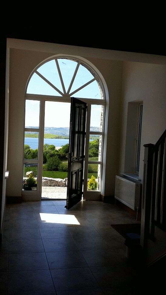 A grand entrance is showcased with an arched doorway leading to a scenic view of Lough Corrib. Natural light streams through large glass panels, illuminating the hallway with tiled flooring. The landscape is framed by greenery, enhancing the inviting atmosphere.