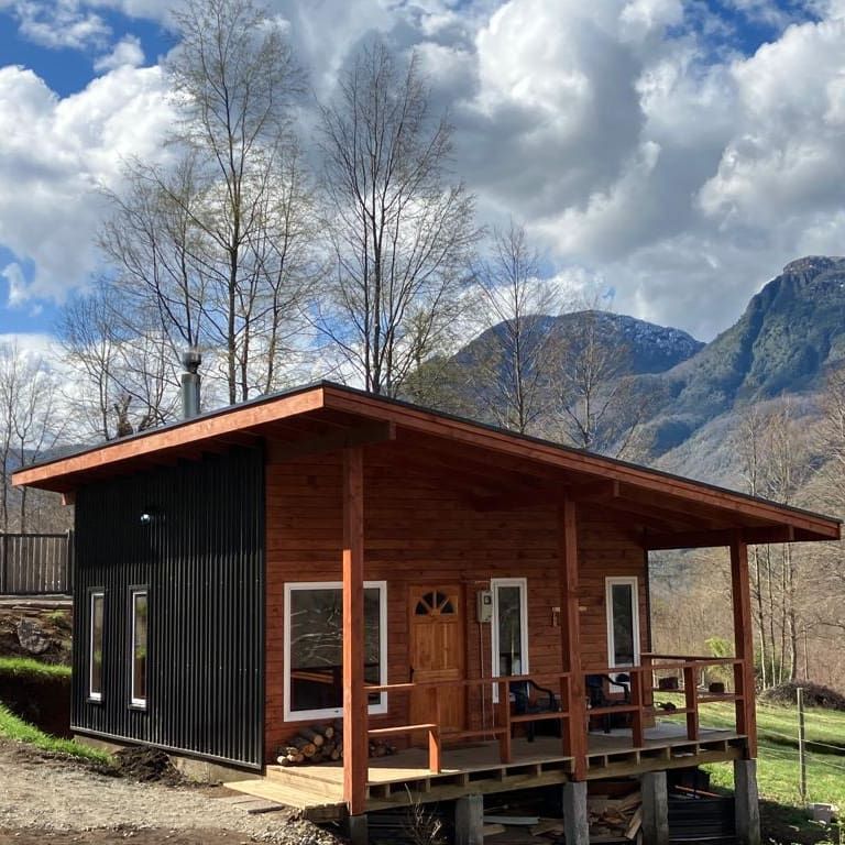 The cabin's exterior showcases a blend of wood and metal elements, situated amidst trees with mountains in the background. A welcoming entrance features a wooden door, while a spacious deck includes seating arrangements, offering a peaceful setting surrounded by nature.