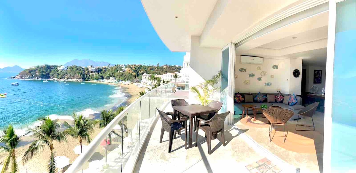 An outdoor terrace displays a dining table surrounded by chairs, offering views of the beach and water. The seating area features colorful cushions and decorative wall art. Palm trees and a clear sky contribute to the inviting atmosphere of this coastal space.