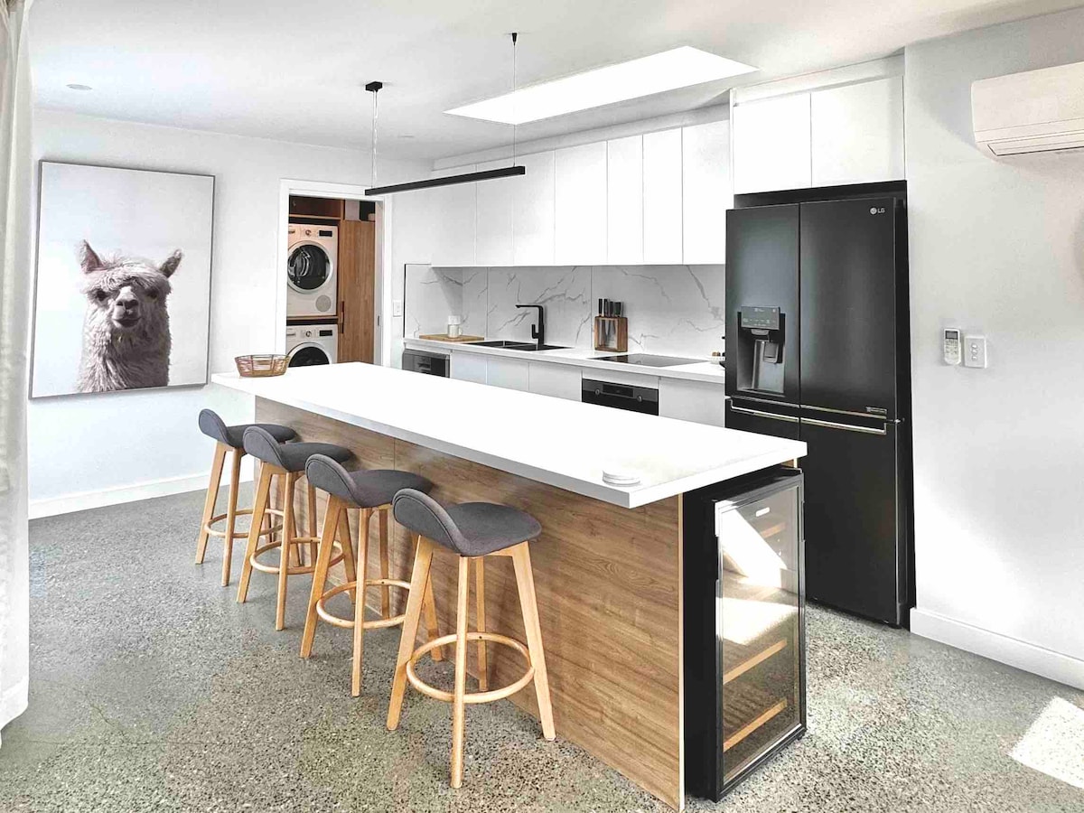 The modern kitchen showcases a spacious island bench with four high stools. Sleek cabinetry and stainless steel appliances complement the polished concrete floor. A large refrigerator and wine cooler add to the functionality, while natural light enters through a skylight above.