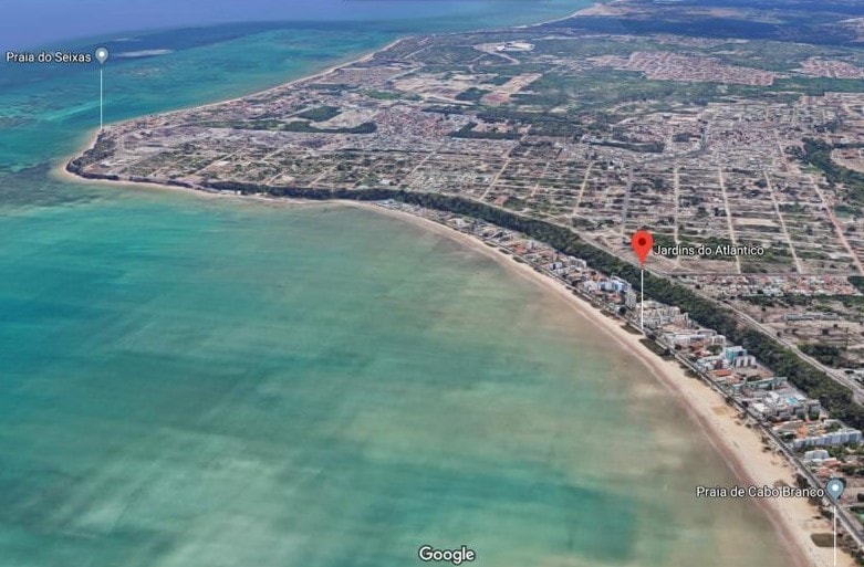 An aerial view captures the coastline and clear turquoise waters bordering a sandy beach. The shoreline is lined with a mix of residential and commercial buildings, showcasing the vibrant atmosphere of the area. Coastal features lead towards undulating land in the distance.