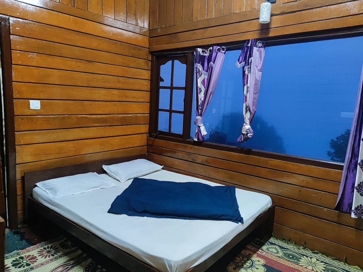 A wooden interior showcases a cozy sleeping area with a spacious bed, featuring white linens and a dark blanket. Large windows allow natural light to filter in, framed by patterned curtains. The floor is covered with a colorful rug, enhancing the room's warmth.