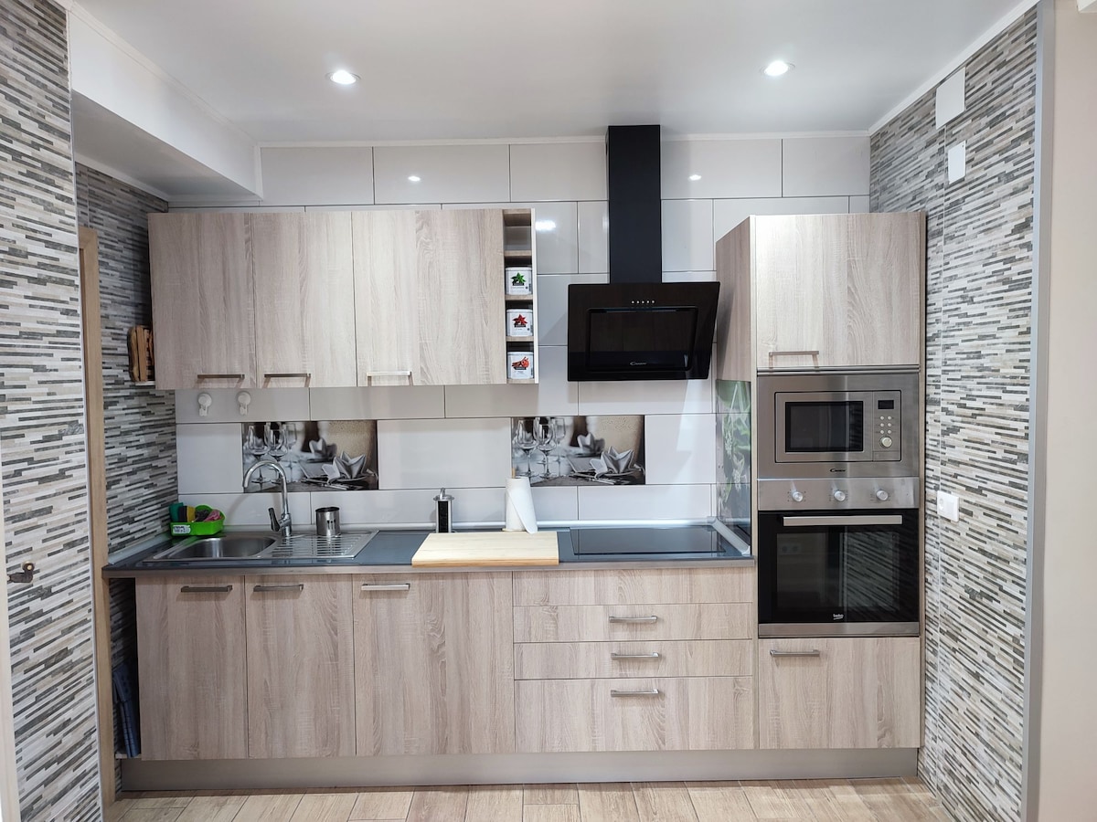 A modern kitchen with light wood cabinetry and a sleek countertop features essential appliances including an oven, microwave, and stovetop. A subtle patterned backsplash complements the design, and natural light enhances the open feel of the space.