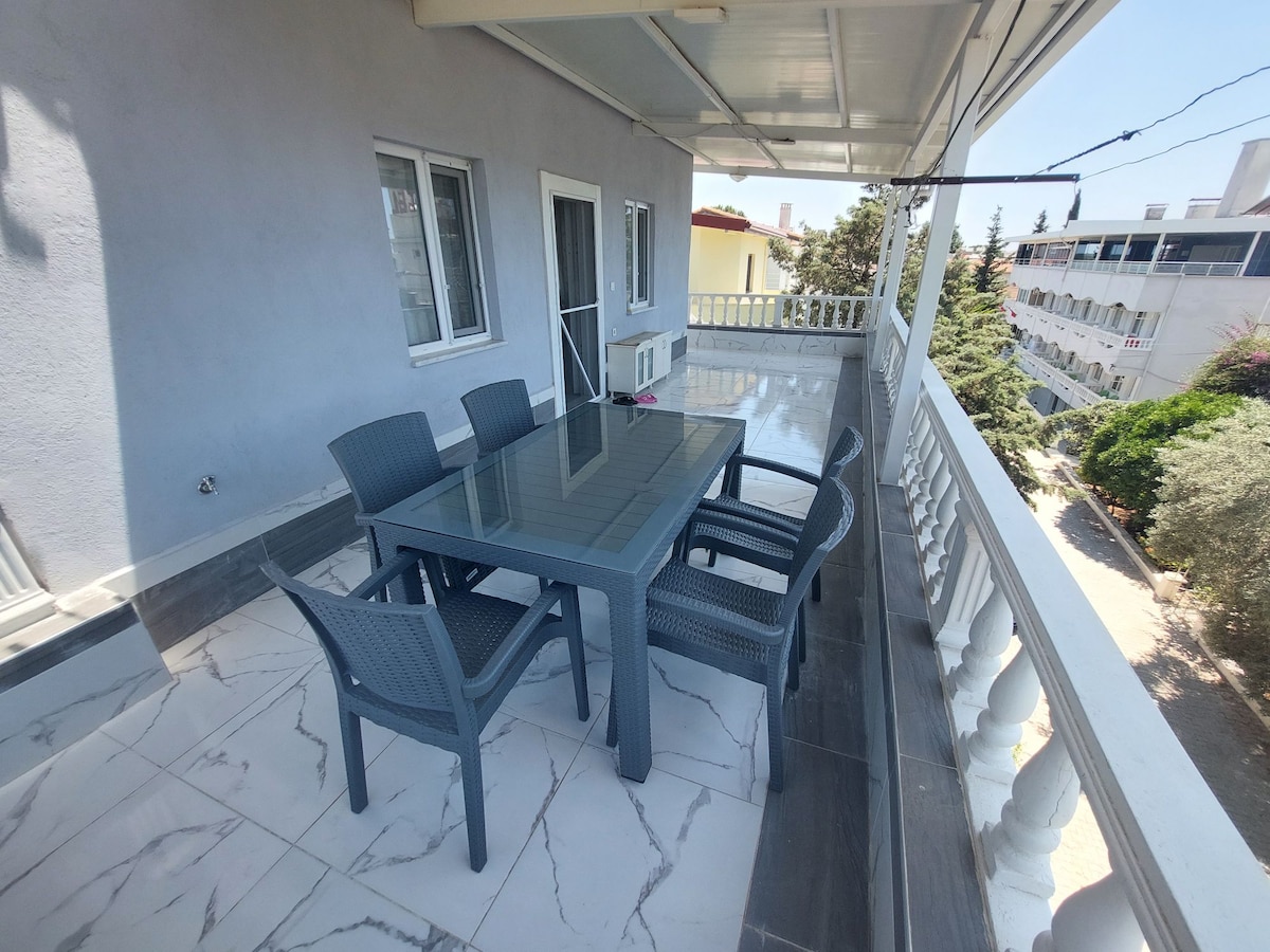 A spacious balcony is furnished with a large table surrounded by six chairs, providing ample seating. The area features a protective overhang, and the tiled flooring offers a modern touch. Views of the surrounding landscape can be enjoyed from this outdoor space.