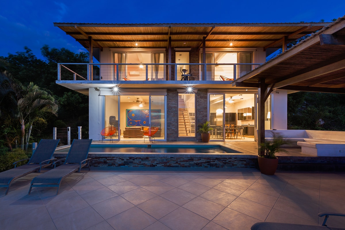 The property façade at dusk reveals a modern design with multiple levels. Large glass doors provide views into the living spaces, while a tranquil pool reflects surrounding lights. Outdoor lounge chairs are positioned near the pool, and a covered area is visible to the right.