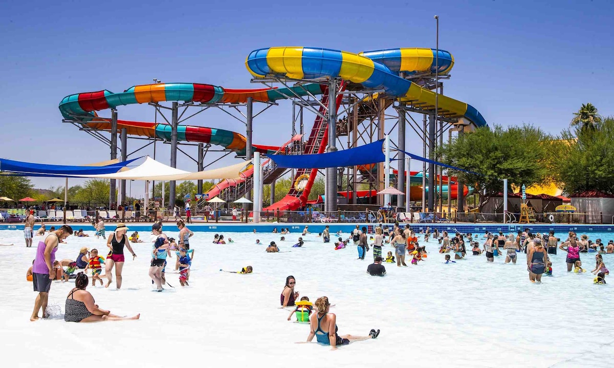 A lively water park scene is captured, featuring colorful water slides in bright yellow, orange, and blue. Guests of various ages are seen enjoying the wave pool, with children splashing in the shallow areas and adults relaxing on the beach-like entrance.